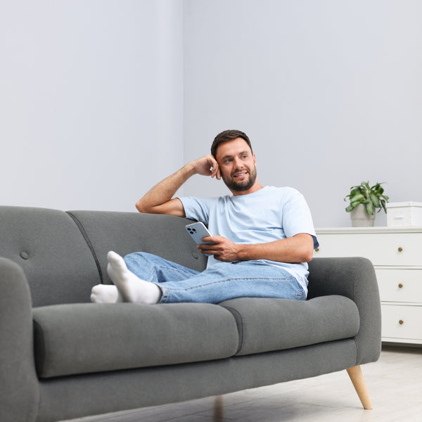 Man sitting on a gray couch holding a phone in a living room.