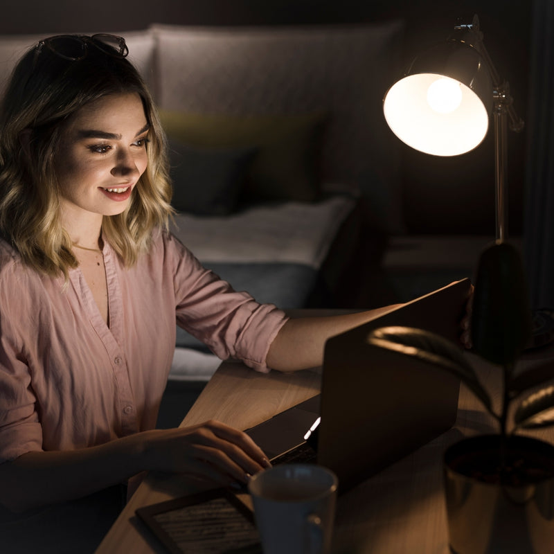 Woman using a laptop in a dimly lit room with a lamp and cup on the table.