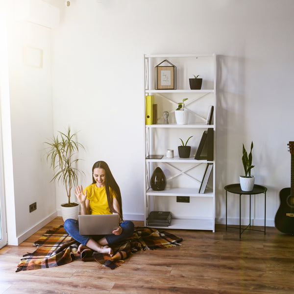 Person sitting on a blanket using a laptop in a room with a bookshelf and plants.